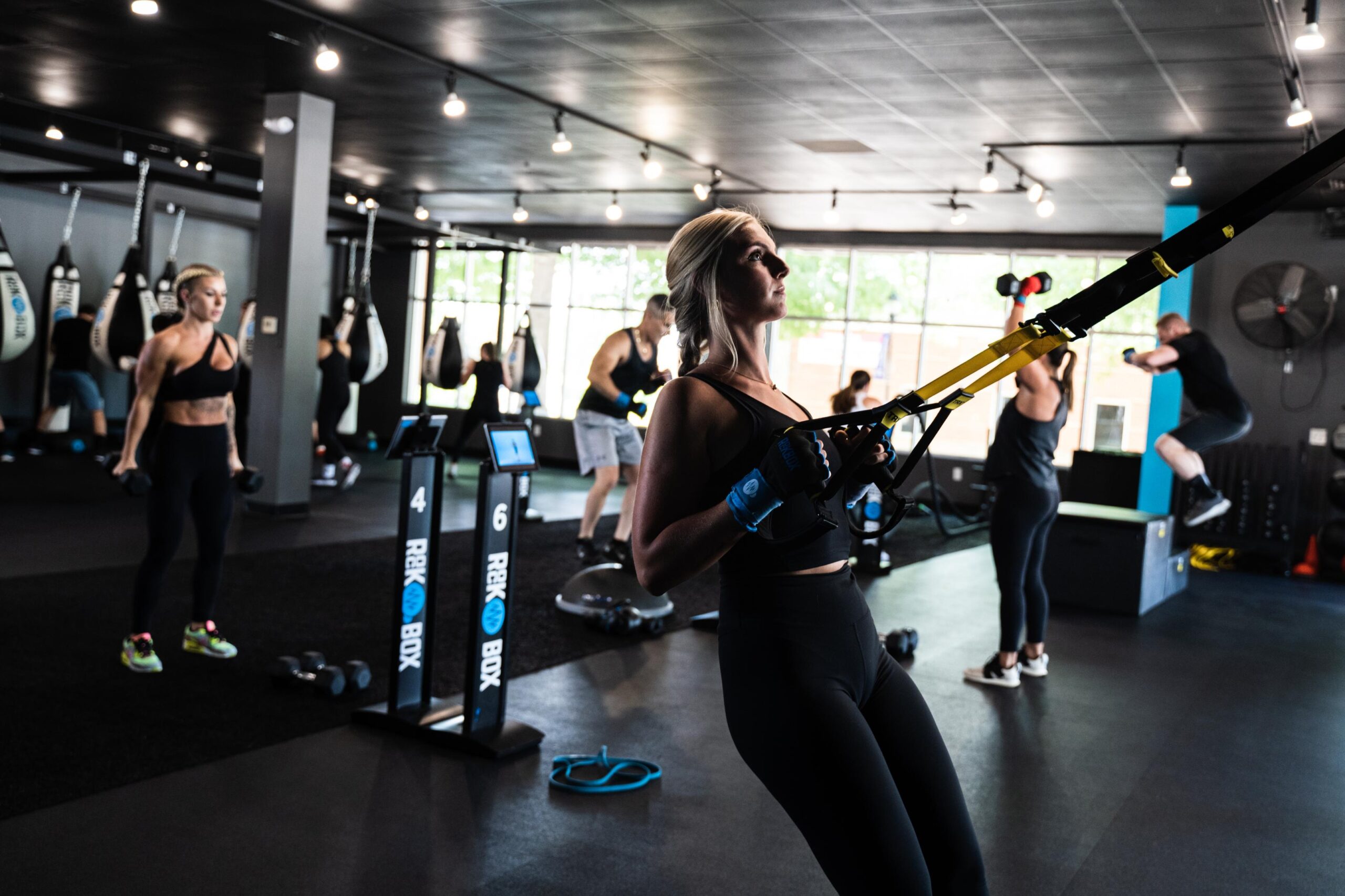 A young woman is using TRX bands in a workout class with other members behind her at stations.