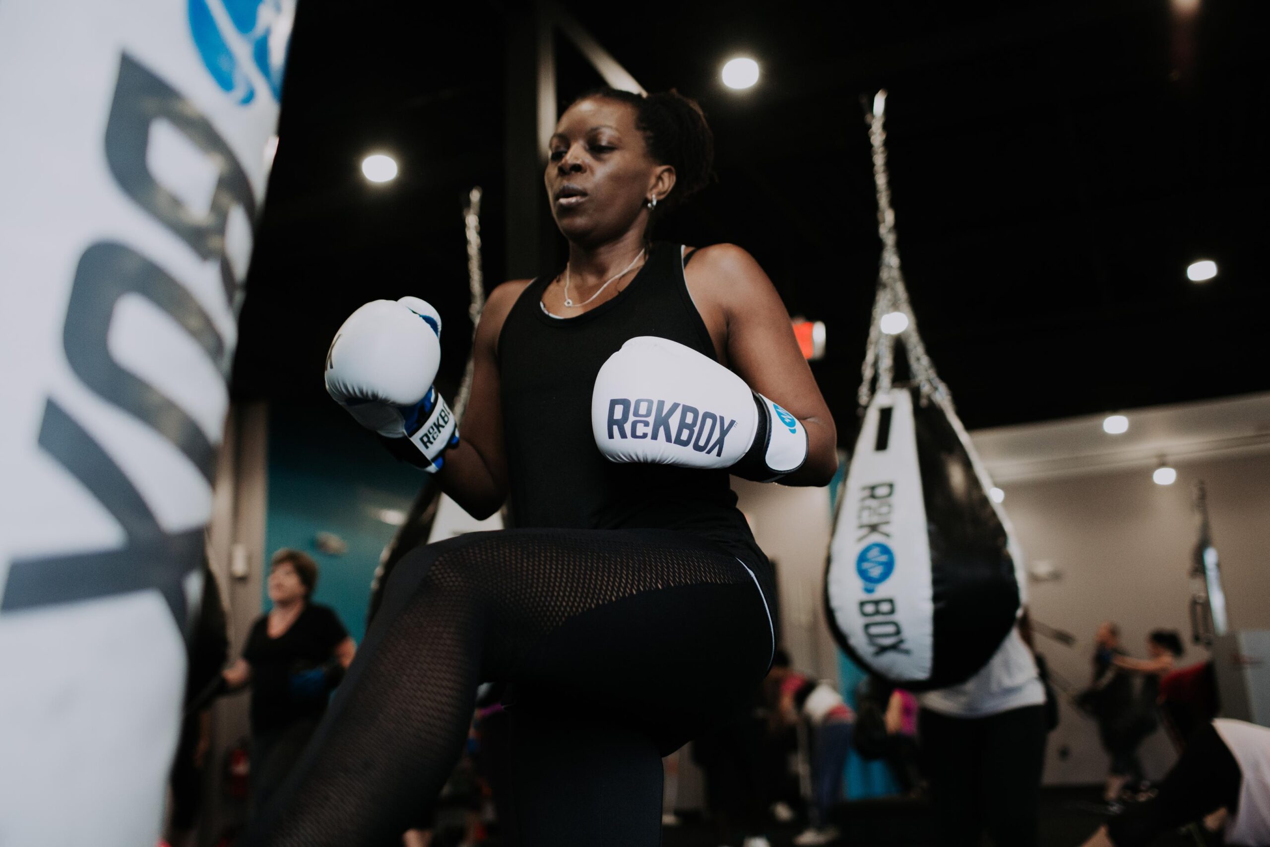 A female wearing boxing gloves prepares to kick the heavyweight bag in a RockBox studio.